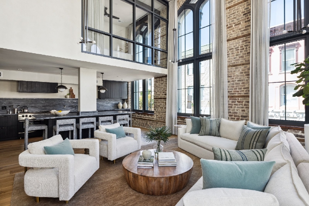 Airy double-height living room with cream seating, pale blue pillows, exposed brick and tall windows furnished by HW Home