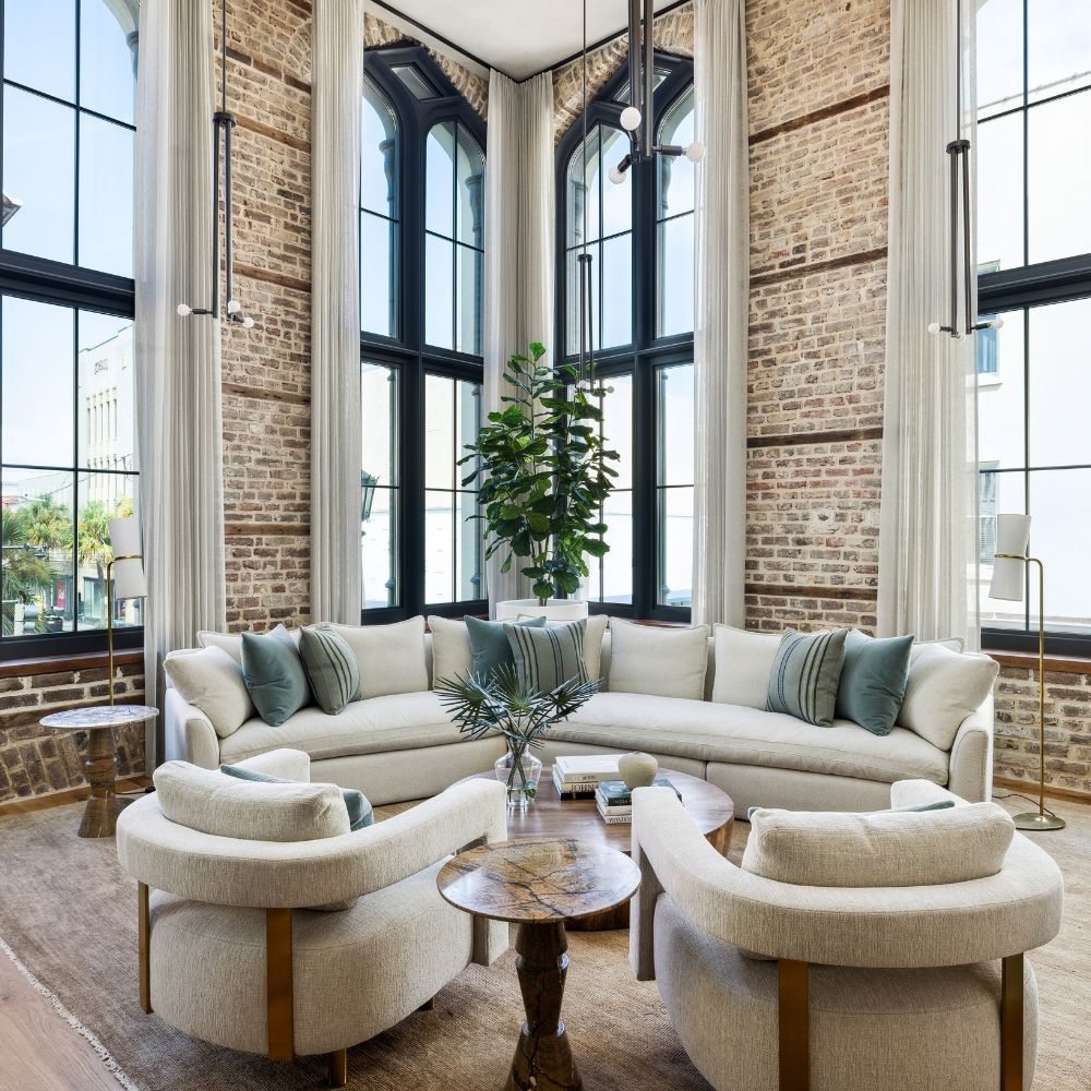 Cream sofas and curved lounge chairs beneath tall windows in a brick-walled living room